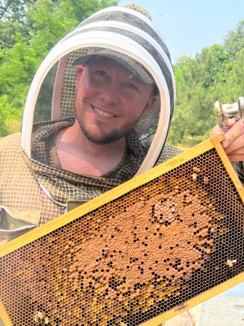Photo of Cory Stevens in a beekeeping suit holding a frame with brood and pollen from a honey bee hive.