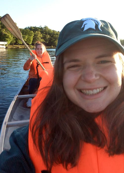 Rachel Drobnak wearing a Spartan ball cap and an orange life jacket is on a canoe with a friend who is paddling.