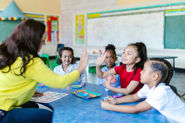 A teacher in a yellow shirt with dark hair high fiving four students at a table.