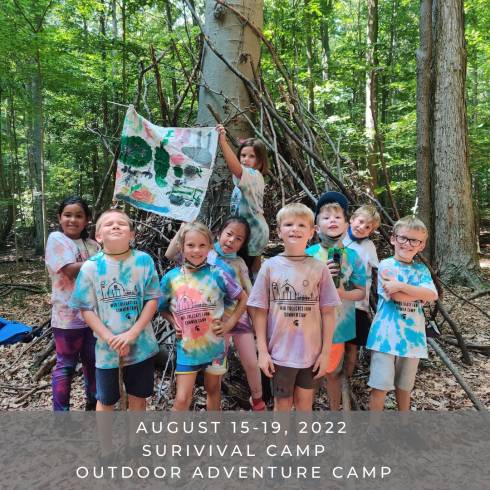 Campers holding their team flag in front of the shelter they made in the woods using sticks and logs.