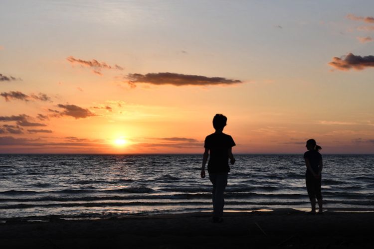 Two people standing on a lake's shoreline with a sunset in the background.