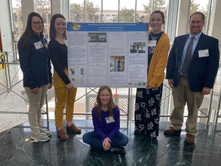 Three students and two adults who participated in the symposium are shown standing and sitting near a display of the student posters that describes their research.