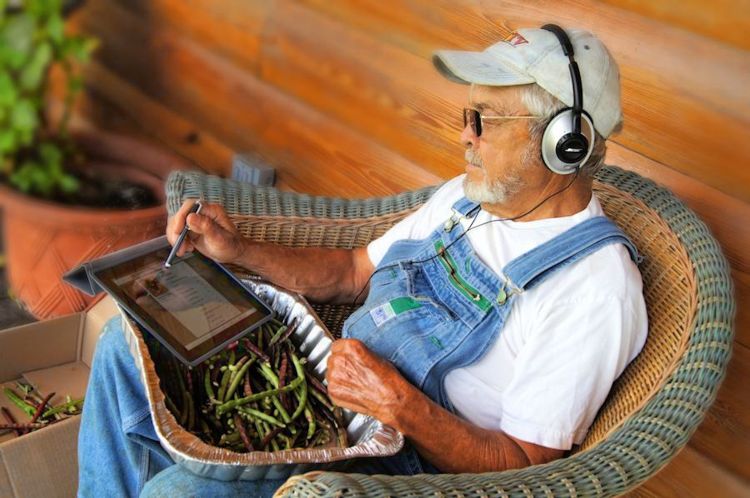 A man sits in a chair with headphones on and an iPad and green beans in his lap.