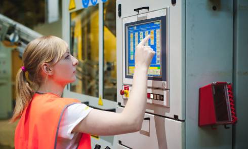 Photo of a woman wearing a safety vest and touching a monitor on a factory machine.