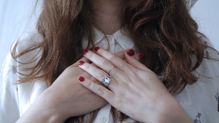 A person with long brown hair and a white shirt holding their hands over their heart.