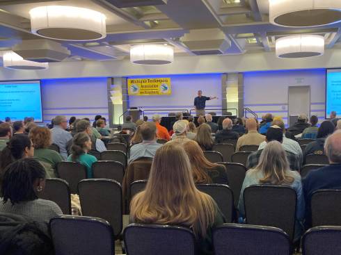 Photo of people sitting in a room facing a presenter and screens.