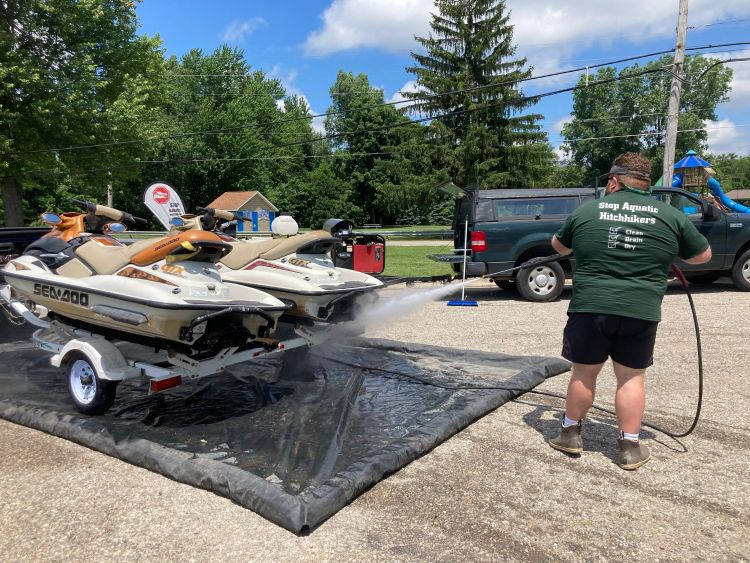 Individual with high temperature and pressure hose washing underside of two jet skis on a trailer.