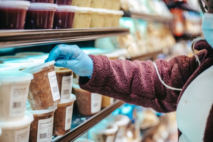 Person with gloves and mask on picking up plastic packaged soup on grocery shelf.