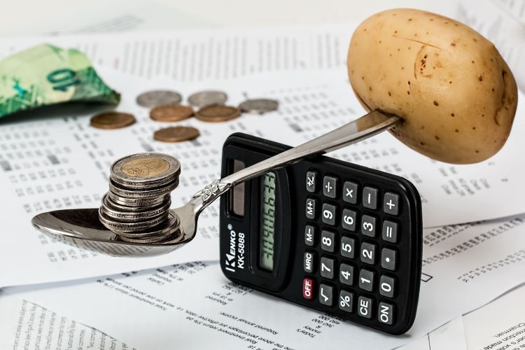 A calculator with a spoon on top of it that is balancing money on one end and a potato on the other.