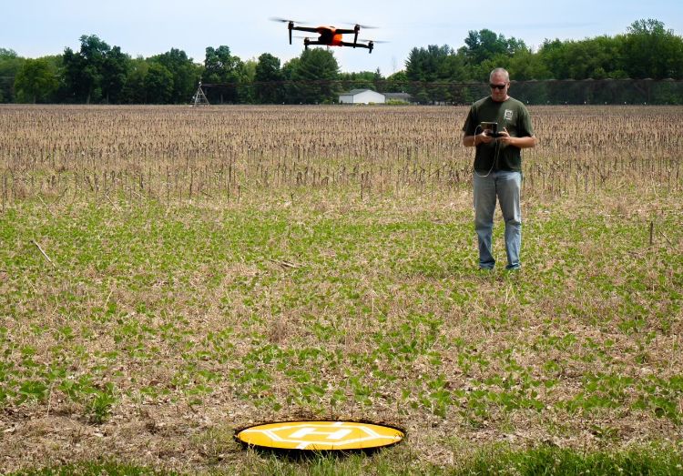 A man stands in a field controlling a drone flying in the air in front of him.