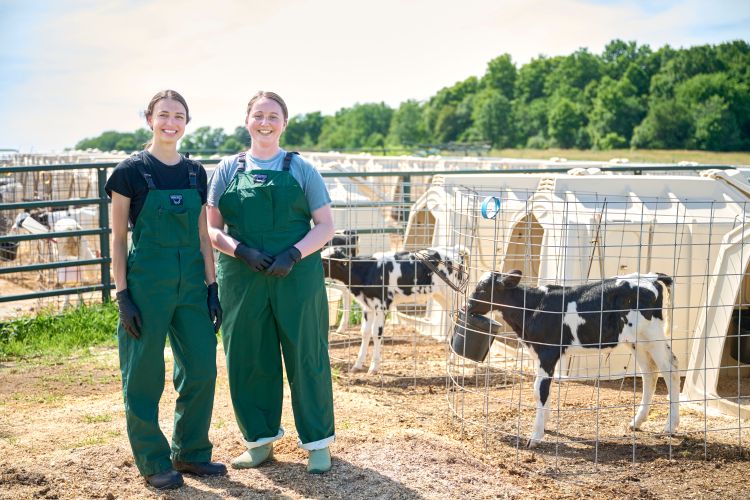 Paiton McDonald and Haylee Reisinger, wearing green bibs, stand in front of Holstein calves in outdoor pens with a blue sky and green trees in the background