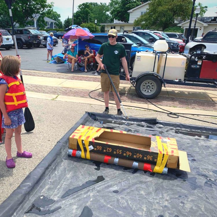 Person standing in parking lot at event demonstrating how to clean boats to stop the spread of aquatic invasive species. Demonstration boat is a small, wooden model on top of a black mat preventing contaminated water from re-entering waterways.