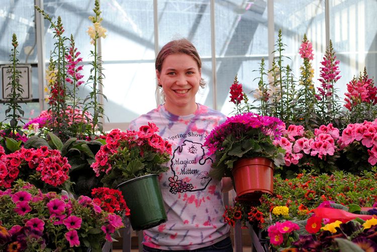 Emma Brinks in a greenhouse holding two flower pots and surrounded by pink flowers.