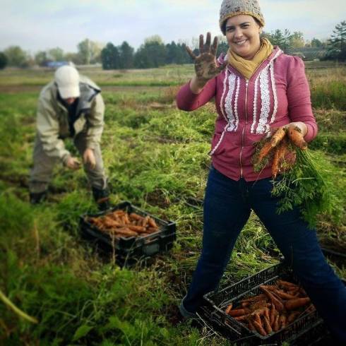 Two people harvesting carrots in a field. One is leaned over, their face obscured by a hat. A woman is in front, stranding wide over a crate of carrots, holding up her hand dirty with soil and smiling big.