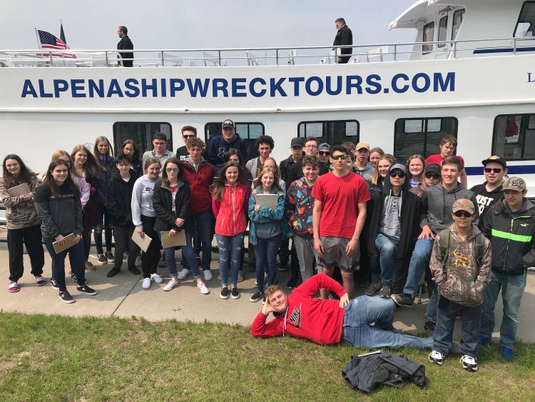 A large group of high school students stand on shore in front of a glass-bottom boat they were going to ride on to explore Lake Huron heritage and habitats as part of their Science in the Sanctuary class. On the boat in the background are the words alpenashipwrecktours.com. Photo: Michelle Cornish, Alpena High School