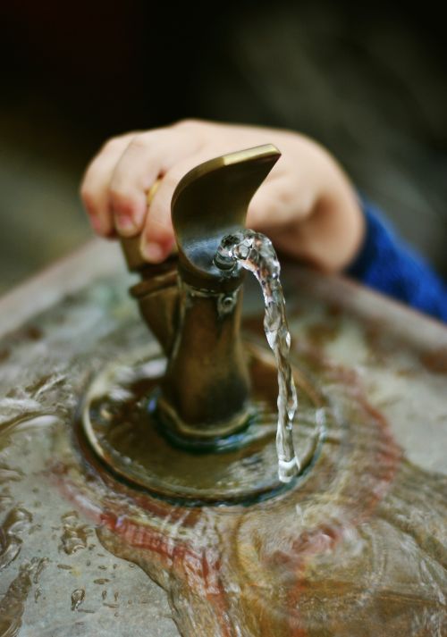 A child's hand is shown pushing down on a drinking fountain handle in order to have water bubble up.