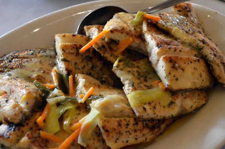 A white serving plate holds several pieces of grilled lake whitefish filets which are covered with seasonings and some vegetables.