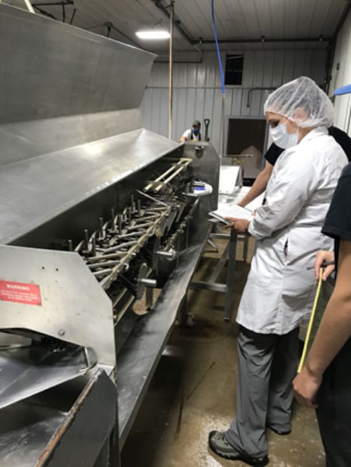 Fish processing plant being inspected by a food inspector who is standing by a machine wearing safety mask and white coat and holding a clipboard.
