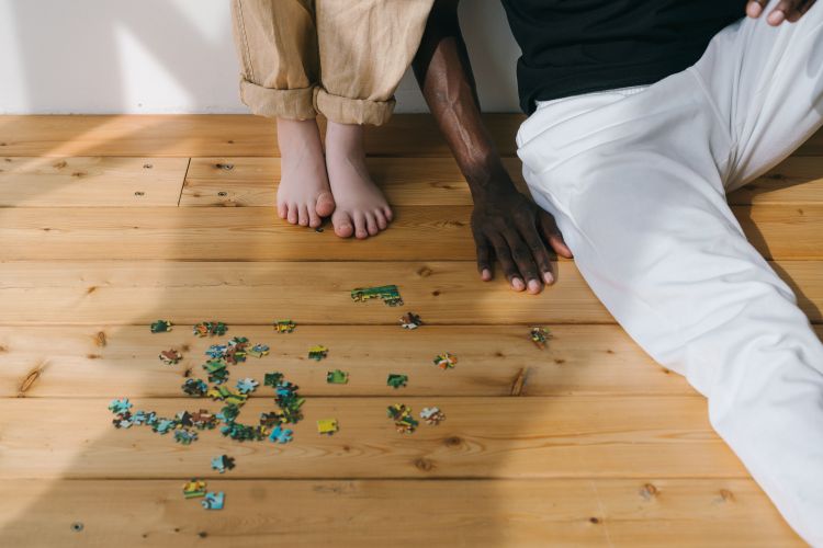 Two people standing and sitting near puzzle pieces on the floor.