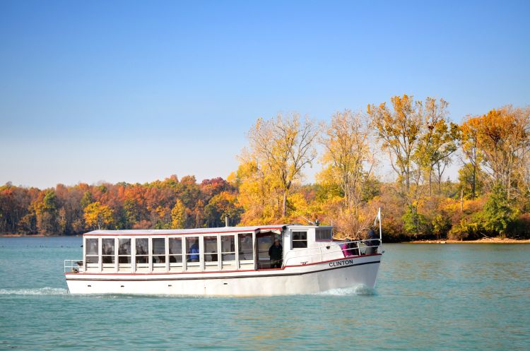 A boat that can seat a large number of passengers under it's cabin is seen on Lake St. Clair. The trees behind are showing fall colors.