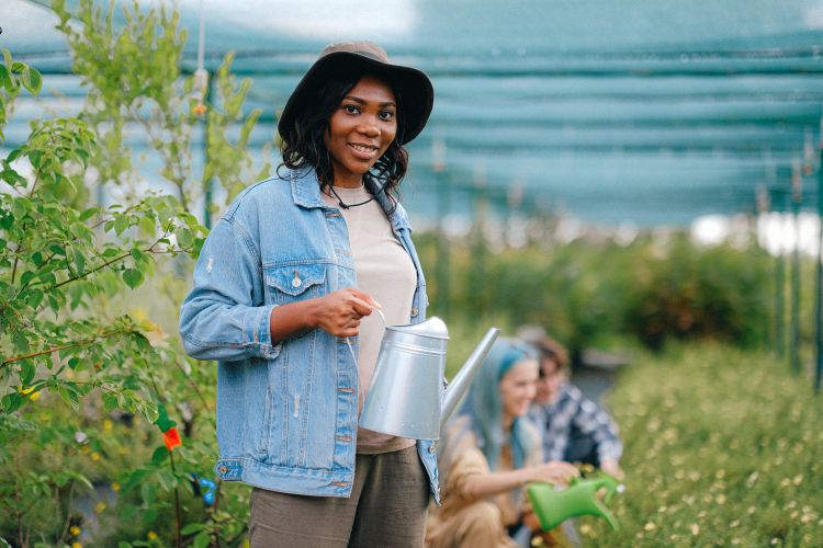 A Black woman holding a watering can. She is wearing a denim jacket and a cowboy hat, and is surrounded by plants covered in a protection net. A few more people watering can be seen in the background.