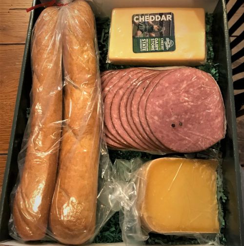 box containing two loaves of bread, one chunk of cheddar cheese, a package of salami and a wedge of gouda cheese