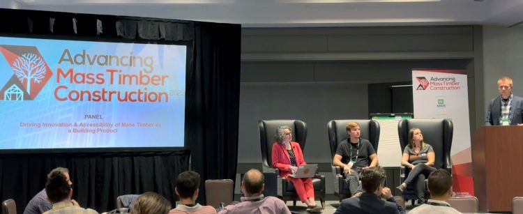 Conference speakers sitting on a stage next to a screen sitting in front of an audience.