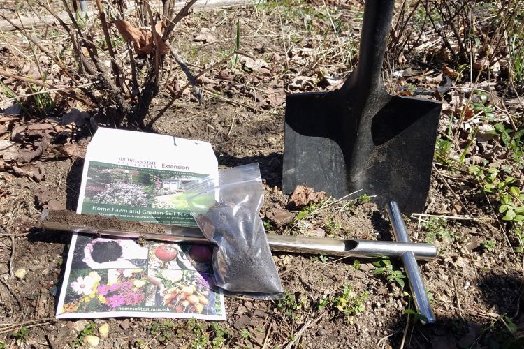 A soil testing probe, shovel, a bag of soil and a soil test kit lay on the ground.