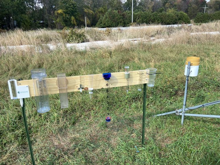 Different types rain gauges attached to a board in a field. These rain gauges ranges from small test tubes to large, complex apparatuses.