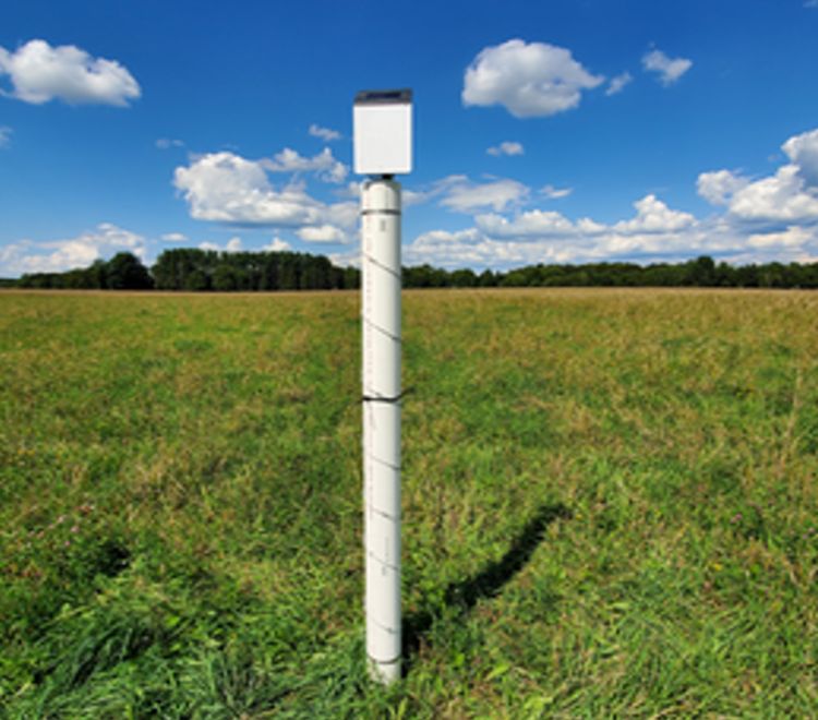 A solar-powered environmental sensor mounted on a tall white pole in the middle of an open grassy field, under a bright blue sky with scattered clouds. Trees line the horizon in the background.