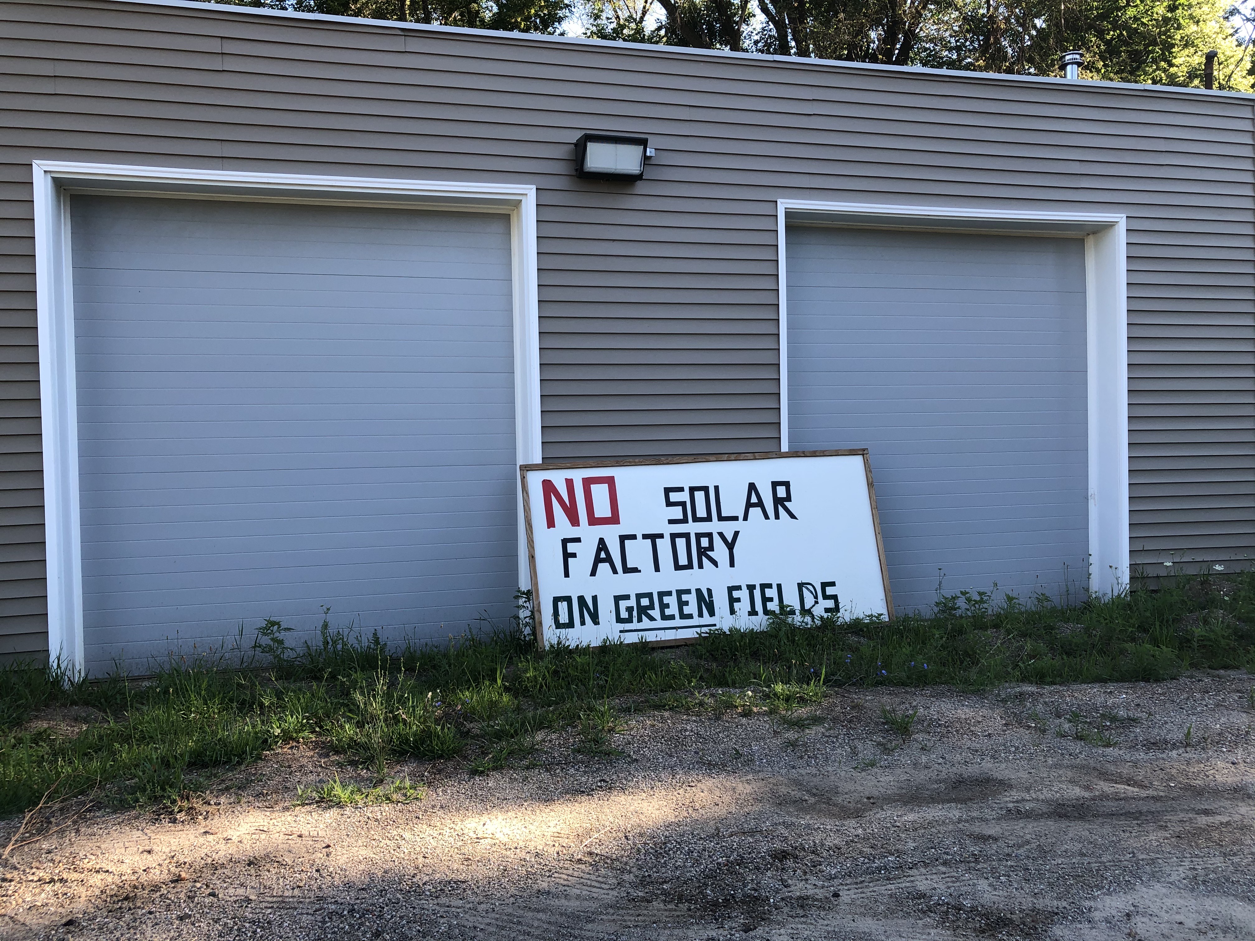 Lettered yard sign saying “No solar factory on green fields” leaning against a beige building.