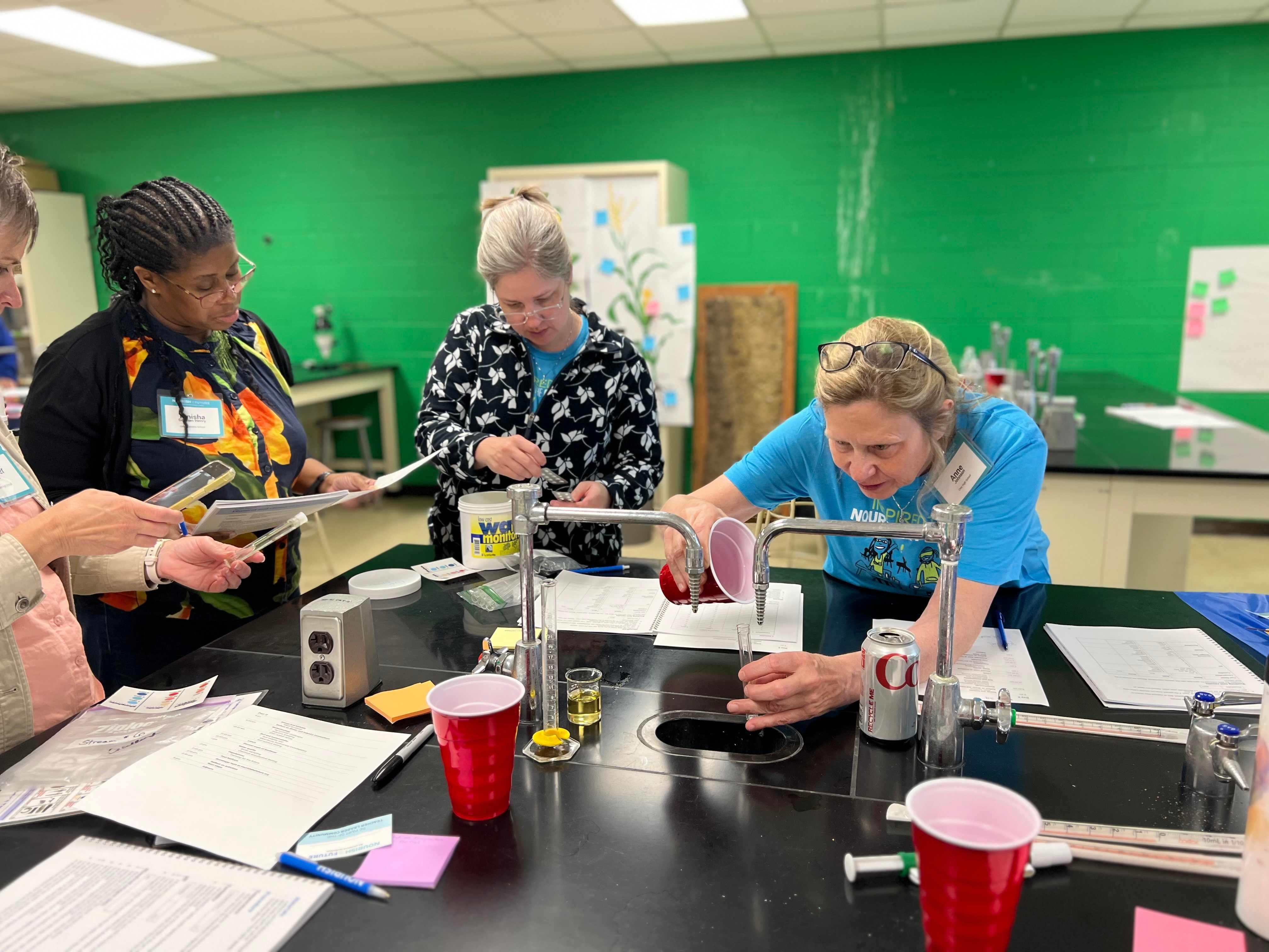 Teachers surround a lab bench cooperating on experiments. 