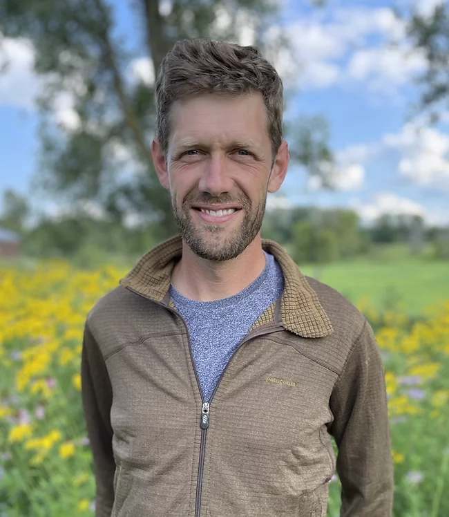 A white man with close cropped beard and hair standing in a field of yellow flowers, wearing a brown zip up, collard shirt.