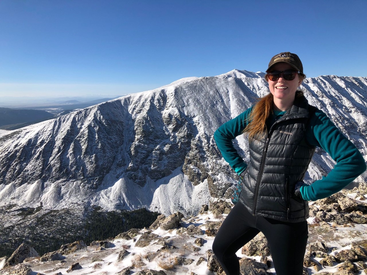 A white woman standing amidst grandiose mountains, wearing sunglasses, a hat, and a green sweater and black puffy vest with her hands on her hips.