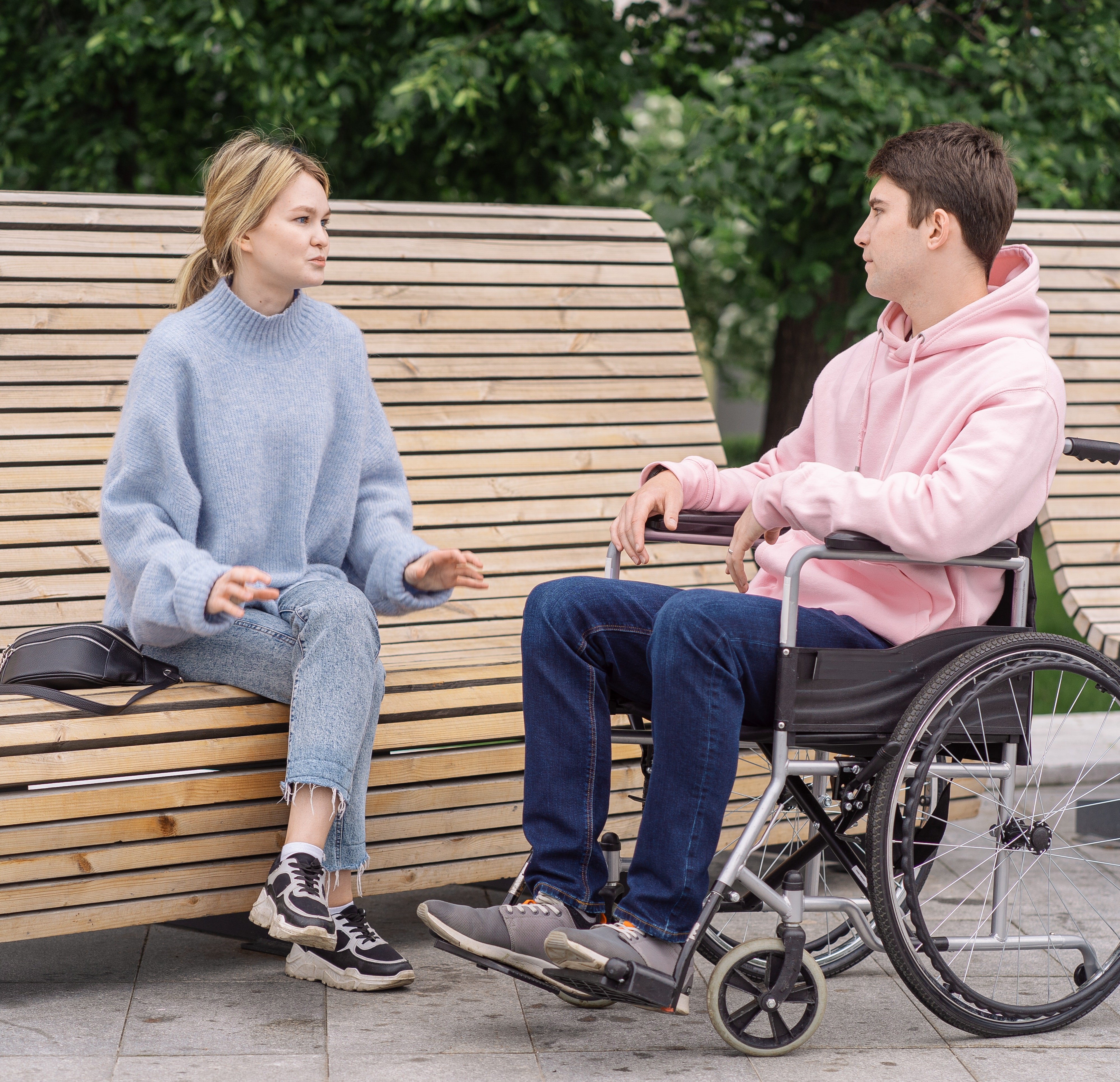 Young man in a wheelchair talking to young woman sitting on a park bench.
