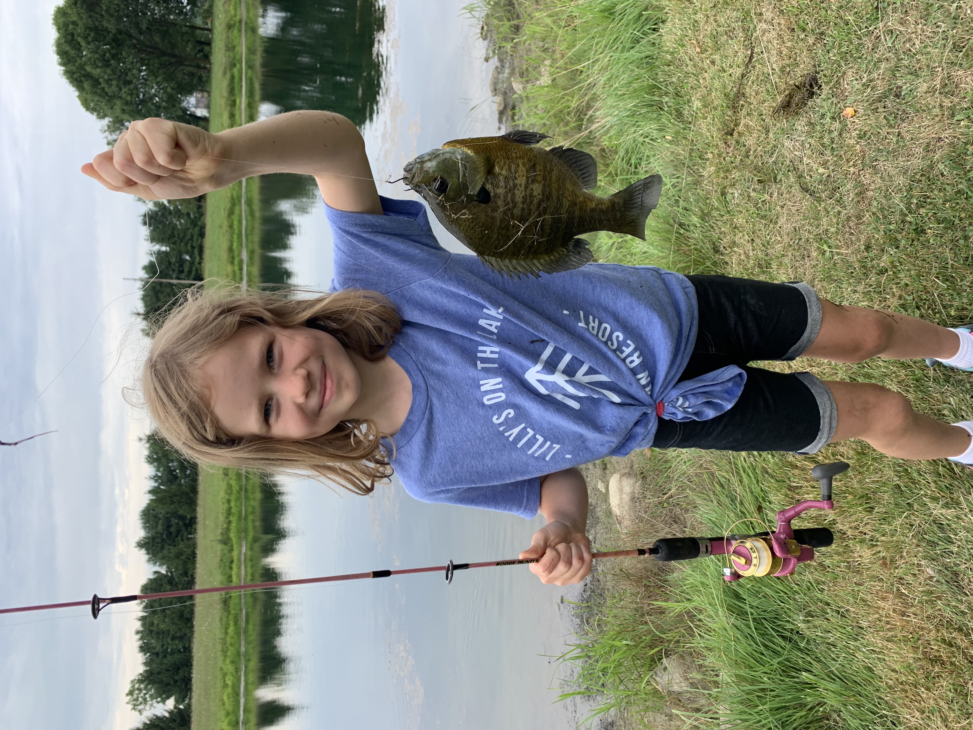 Lilly holding up a fish and fishing rod near a lake