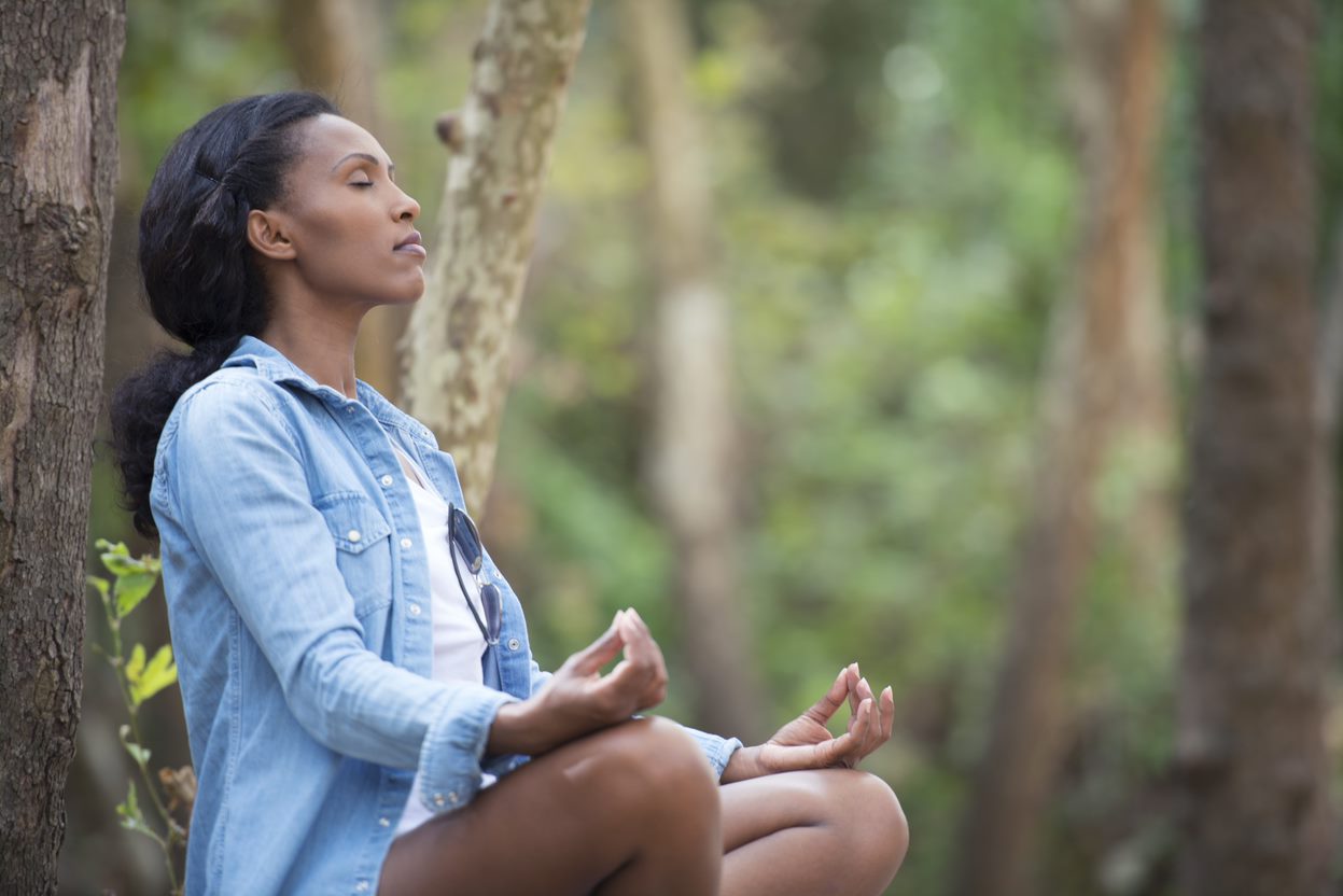 A woman sits against a tree peacefully with her eyes closed.